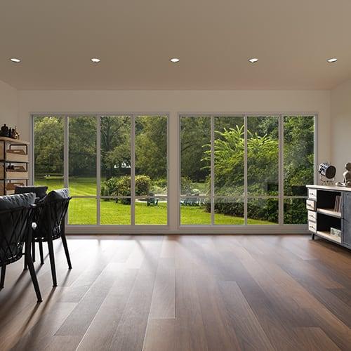 A florida room, inside room view that shows hardwood floors, black chairs, and large windows with view that shows a yard with small bushes and grass.